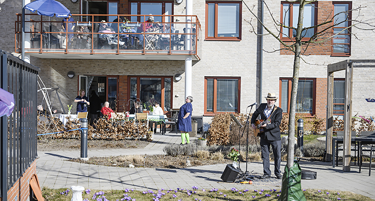 En man med gitarr och mikrofon spelar. De gamla sitter ovanför på en balkong. Solen skiner.