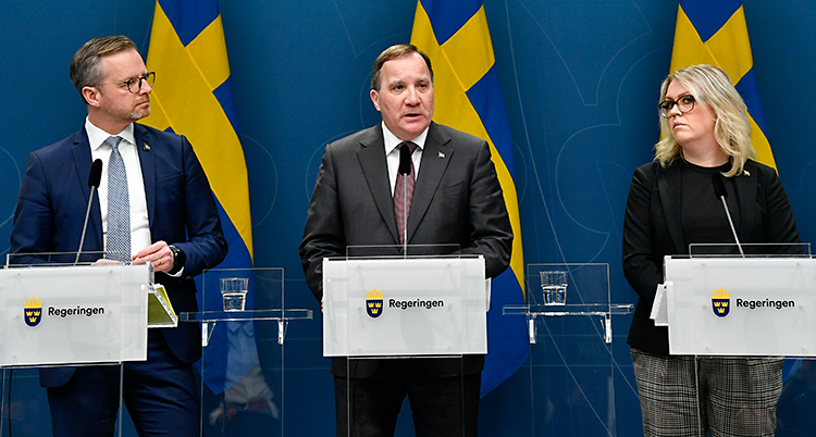Tre politiker står på en scen. De talar i mikrofoner. I bakgrunden syns svenska flaggor.