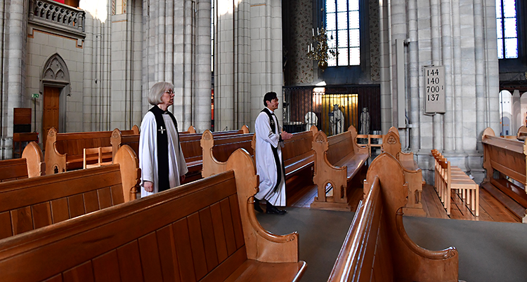 Tomma bänka i kyrkan. Två präster i vita kläder går genom kyrkan