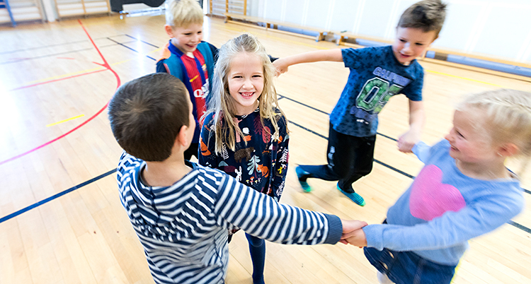 Fem barn är på ett gymnastikgolv. Fyra av barnen håller varandra i hand i en ring. De springer runt. I mitten står det femte barnet och ler mot kameran.