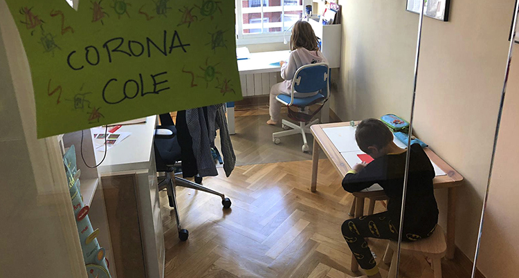 två barn sitter inne framför varsin bänk och gör läxor. Vi ser dem fotade uppifrån. Snett i förgrunden hänger en lapp där det står något om corona på spanska.