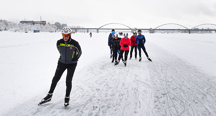 En istäckte av tunn snö. I bakgrunden är en bro. En grupp med skridskoåkare.