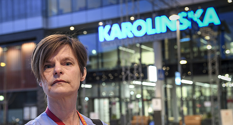 Hon har brunt kort hår och tittar in i kameran. Hon står framför ett sjukhus.