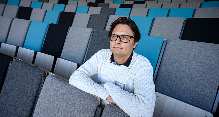 Han sitter på en läktare. Stolarna är tomma runt honom.