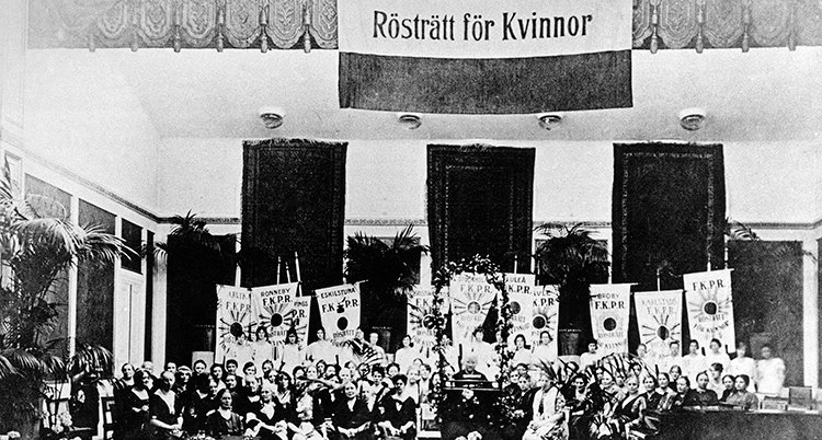 En gammal svartvit bild. Kvinnorna sitter på rad under en skylt med texten Rösträtt för kvinnor