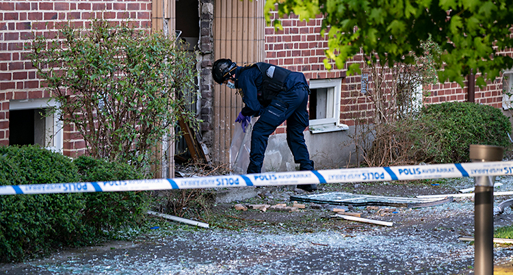 Bilden är tagen utomhus. Vi ser ett hus med lägenheter. Poliserna har spärrat av med band. Innanför bandet jobbar en polis vid porten. Porten har blivit trasig och det ligger skräp från explosionen på marken.