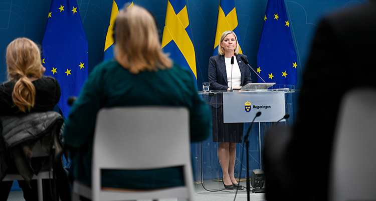 Hon står och pratar vid en talarstol. Hon pratar i en mikrofon. Bakom henne finns svenska flaggor. Framför sitter journalister på stolar.
