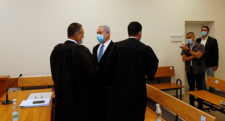 Netanyahu är i ett rum i en domstol. Han har på sig kostym. Han har också ett skydd för munnen. Det beror på viruset corona.
