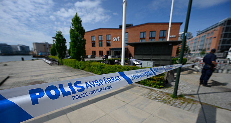 Vi ser ett blått och vitt band där det står polis. I bakgrunden är ett stort hus.