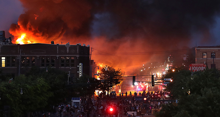 massor av människor på en gata i minneapolis. Bilden är tagen från långt håll. Det är mörkt men längre bort i horisonten står en byggnad i brand och kastar ett rött eldsken på dem som protesterar.