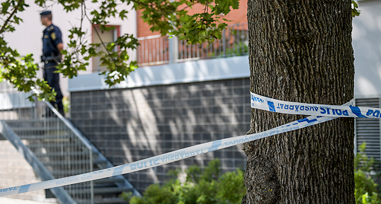 Bilden är utanför ett hus med lägenheter. Poliser har spärrat av med band runt ett träd. I bakgrunden står en polis.