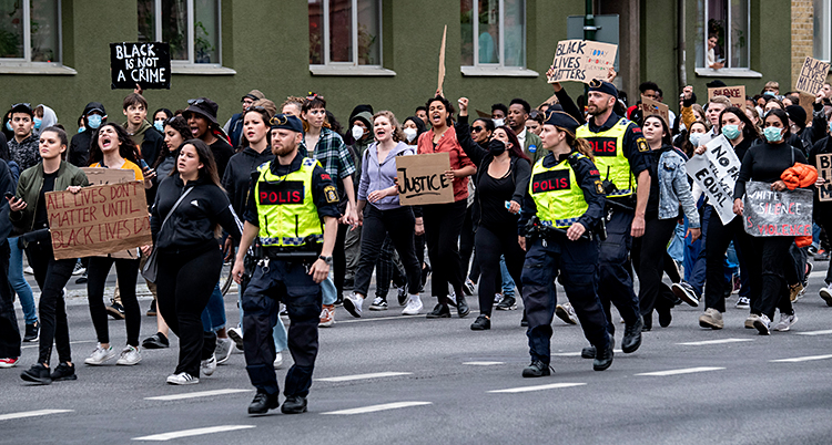 Många människor går på en gata. De har skyltar där det står saker. Vid sidan av dem går några poliser.