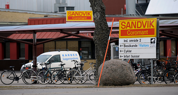 Bilden är tagen på gatan utanför företaget Sandvik. Vi ser en skylt där det står Sandvik. Vi ser också en vit bil som står framför företagets stora byggnad.