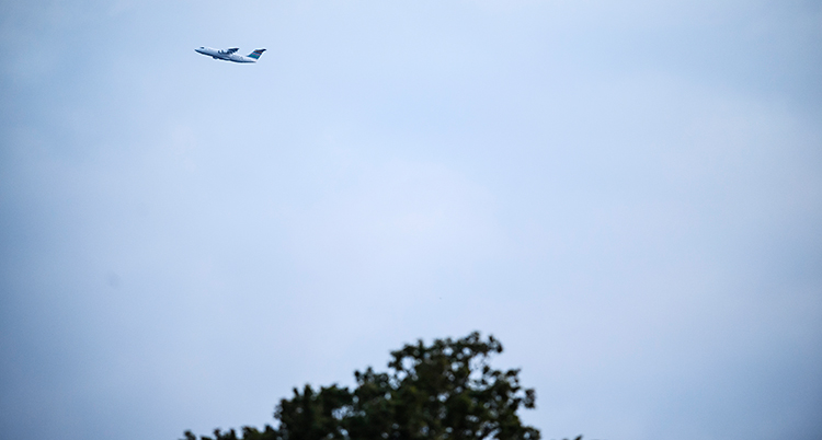 Vi ser ett flygplan uppe i luften. I nedre delen av bilden syns toppen av träd.