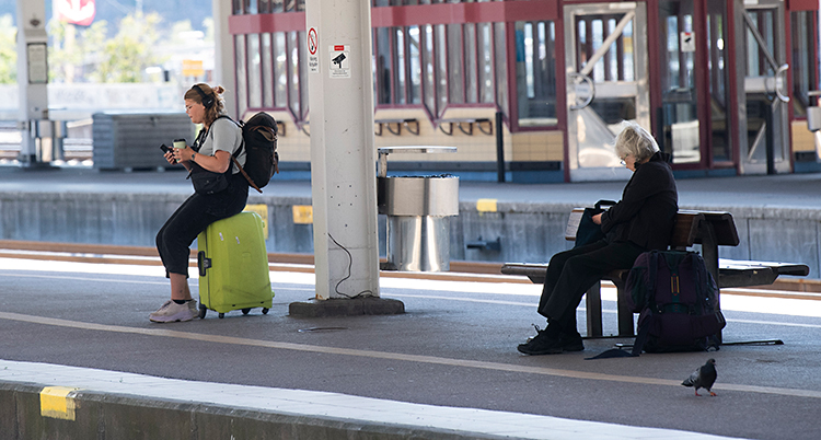 Vi ser en perrong på en station för tåg. En ung kvinna sitter på en väska. En äldre kvinna sitter på en bänk.