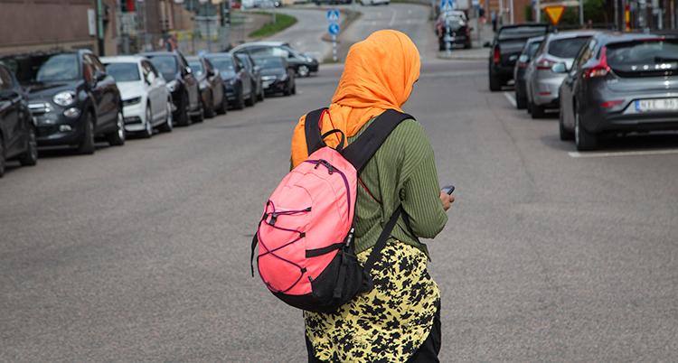 En kvinna med orange slöja och ryggsäck tittar på sin mobil. Hon har ryggen mot kameran.