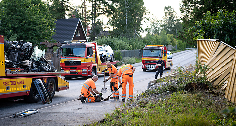 Bilden visar en väg i ett område med villor. Ett staket är trasigt. Några personer i orange kläder städar på vägen. Där finns också två trasiga bilar som står på bärgningsbilar.