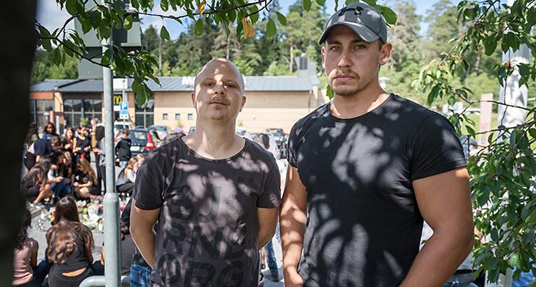 Bilden är tagen utomhus. Två män står och tittar allvarligt in i kameran. I bakgrunden syns en massa ungdomar som har samlats för att sörja.