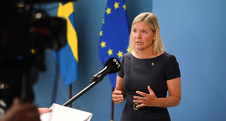 Hon står i ett rum och pratar i en mikrofon. En kamera filmar henne. Hon har blont hår. I bakgrunden är en blå vägg. Vid väggen finns Sveriges flagga och EUs flagga.