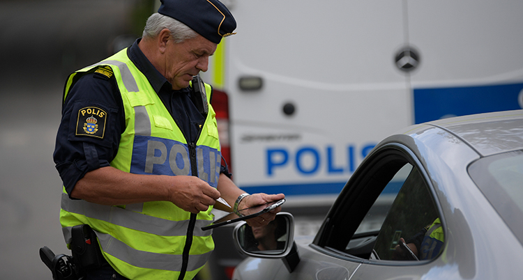 en polis i gul väst tittar på ett körkort. Han står vid en bil som har bliivit stoppad. Förarens ruta är nedvevad.