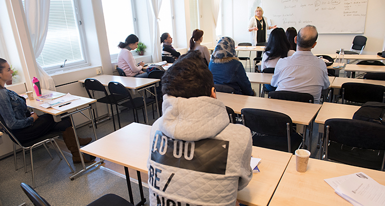 Vi ser ett klassrum. En lärare står vid tavlan och pratar. Elever sitter i bänkar och lyssnar.