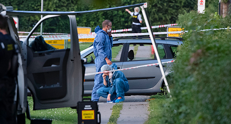 Bilden är tagen utomhus. Två personer har på sig skyddskläder. De står vid en bil. Runt om har poliser spärrat av med band.
