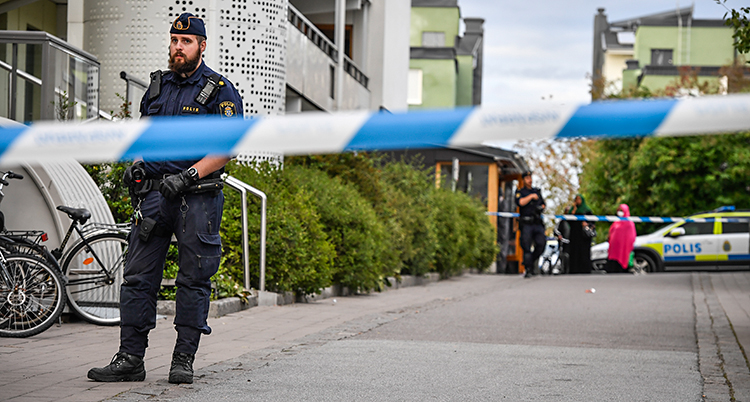Bilden är tagen utomhus. Vi ser hus med lägenheter. Poliser har satt upp blå och vita band. Två poliser står innanför banden. Längst bort syns en polisbil.