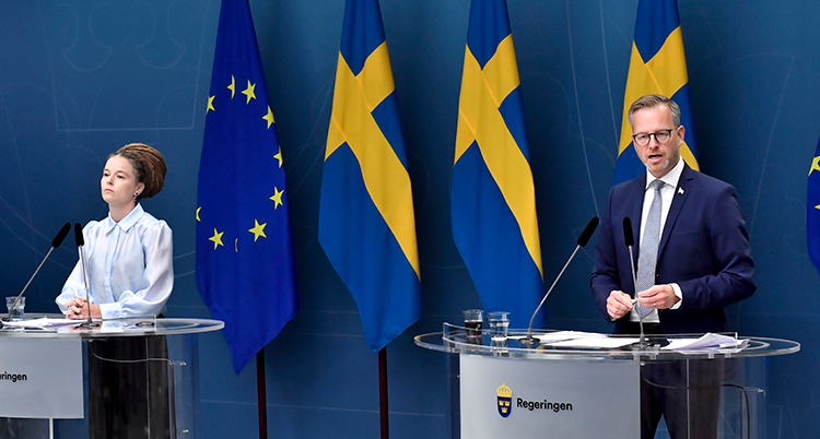 Lind och Damberg står i varsin talarstol en bit från varandr. Bakom dem är tre svenska flaggor påg en EU-flagga.