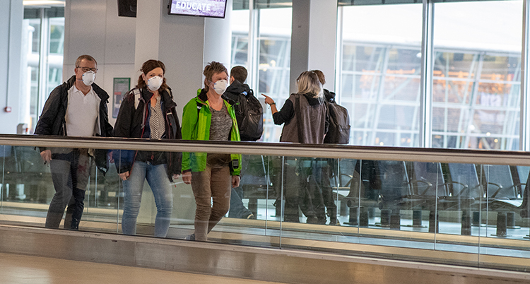 Tre personer har munskydd. De åker på ett rullband på flygplatsen. Bakom dem syns stora fönster.