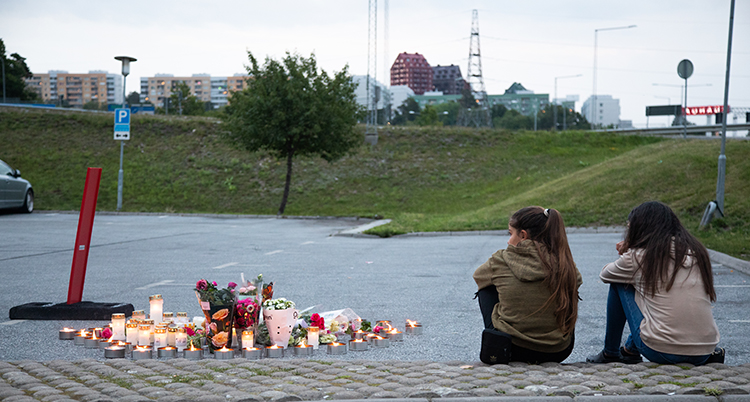 Två flickor siter med ryggen mot kameran. Bredvid dem brinner an massa ljus och där ligger många blommor på marken.