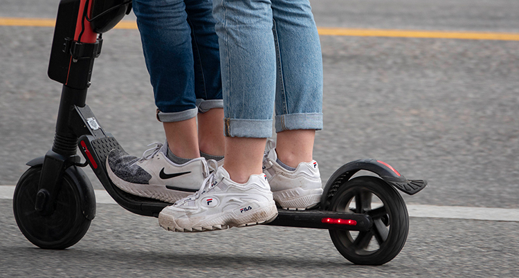 En bild på två personer som åker sparkcykel. Bilden visar bara deras ben och fötter.