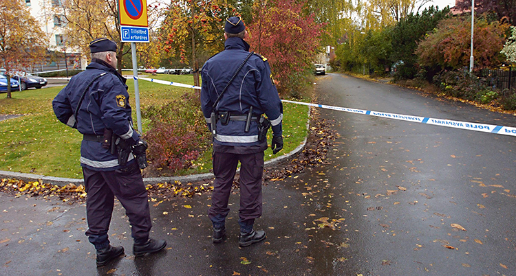 Bilden är tagen utomhus. Det är höst och regnigt. Två poliser står på en gata. De har ryggen mot kameran. Poliserna har spärrat av gatan med blå och vita band.