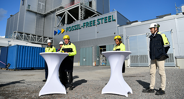 Bilden är tagen utomhus. Flera personer står utanför en fabrik. De har på sig skyddshjälmar.