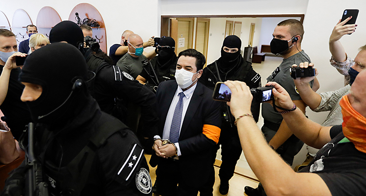 En man är i en domstol. Han går genom ett rum. Han har kostym, munskydd och handbojor. Poliser med masker och stora gevär följer honom. Runt om finns journalister som filmar och tar bilder.