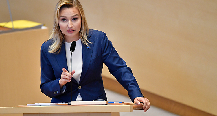 Ebba Busch står i talarstolen i riksdagen. Hon pratar i en mikrofon. Hon har vit tröja och blå kavaj.