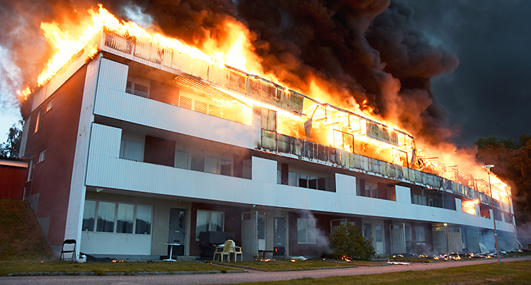 Vi ser ett stort hus med lägenheter. Huset är fyra våningar. I toppen av huset brinner det kraftigt. Det kommer mörk rök från elden.