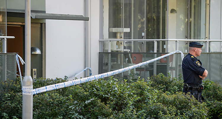 Vi ser ett hus med lägenheter. Poliserna har spärrat av med band som är blå och vita. En polis står och vaktar.