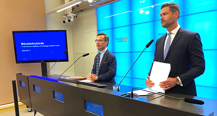 Två män står vid en bänk. De pratar i varsin mikrofon. Båda männen har kostym och slips. Mannen till vänster har glasögon. Bakom dem på väggen står det Sveriges riksdag.