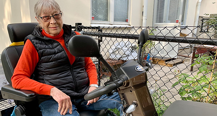 Hon sitter i sin permobil. Hon har grått hår och glasögon. Hon har en röd tröja och en svart väst. Hon tittar in i kameran.