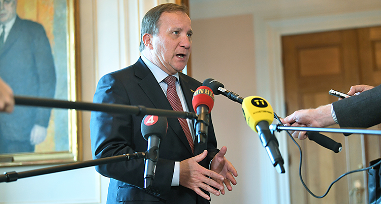 Stefan Löfven står och pratar. Han har kostym och slips. Journalister sträcker fram mikrofoner på långa pinnar.