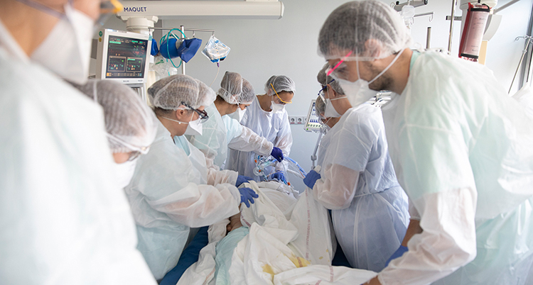 Flera personer står runt patienten som ligger i en säng. Patienten får andas i en mask. Alla har munskydd över munnen.