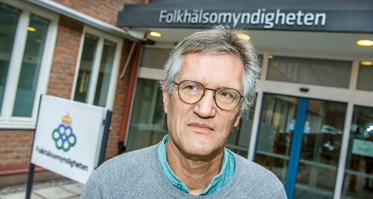 Han har glasögon och grå tröja på sig. Han står framför ett hus. Det står Folkhälsomyndigheten på en skylt bakom honom.