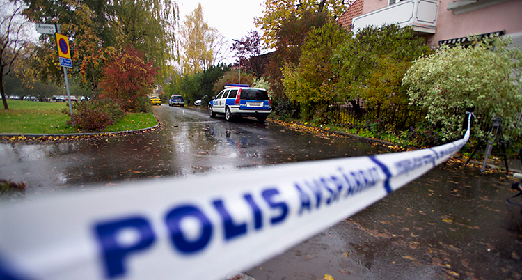 Bilden är tagen utomhus, på en gata. Det är höst. Det är blött på asfalten och trädens löv har olika färger. Poliserna har spärrat av med ett band. Bakom bandet står en polisbil.