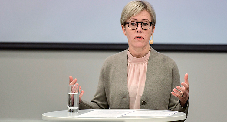 Hon pratar till journalister. Hon står vid ett bord. Hon har kort ljust hår och glasögon.