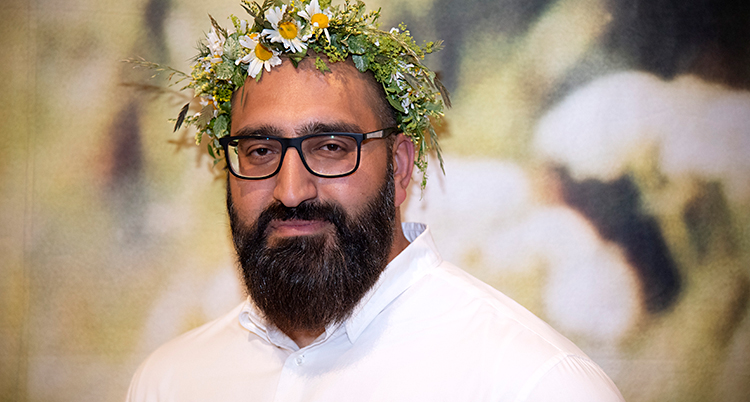 Han har en krans av blommor på håret. Han har glasögon och skägg.