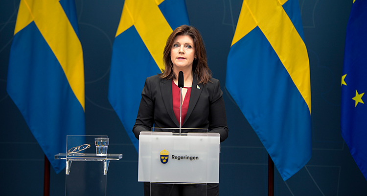 Hon står på en scen och talar. Sveriges flagga syns bakom henne.