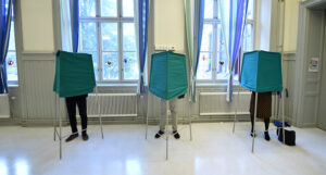 tre personer står bakom tre röstbås i en sal. Vi ser bara deras ben.