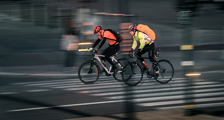 Två cyklister cyklar bredvid varandra. De har hjälmar på sig. De cyklar snabbt. Bakgrunden är suddig. Det är kväll på bilden.