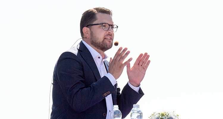 En gammal bild på Åkesson. Han har en mikron vid sin mun och han håller upp händerna. Han har kostym på sig.