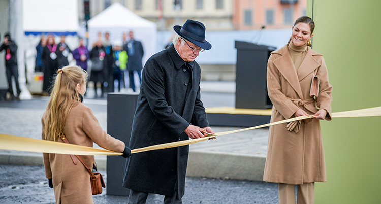 Kungen håller i en sax. Han klipper av ett band som är guldfärgat. Kungen bär hatt. På varsin sida om honom står kronprinsessan och prinsessan Estelle. De har båda beigea kappor på sig.